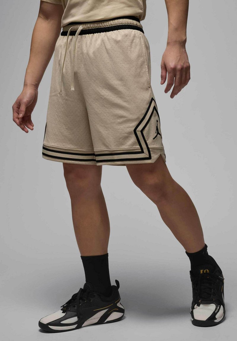 Pantalones cortos deportivos beige con acabado texturizado, acentos y rayas negras, que presentan un logo. Combinados con zapatillas negras y blancas.