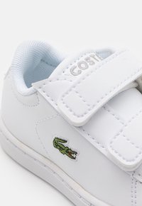 Deportiva de cuero blanca con tiras de velcro, cuello acolchado y forro de malla azul. Presenta un logo de cocodrilo bordado en verde. Diseño sencillo.