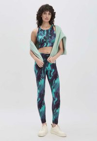 Braagtop en hooggetailleerde legging van rekbaar materiaal, met een blauw en paars abstract patroon. Model houdt een lichtgroene trui vast.