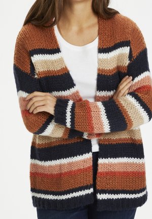 Person trägt eine dicke, mehrfarbig gestreifte Strickjacke über einem weißen Hemd, die Arme verschränkt, mit Rost-, Marine-, Beige- und Weißtönen.