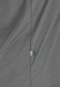 Tessuto grigio con piccolo motivo a quadrati e una piccola etichetta blu con una piccola croce scandinava gialla cucita nella cucitura.