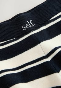 Primer plano de la cinturilla de punto con rayas blancas y negras, con borde acanalado y texto bordado en blanco "self" en la banda.