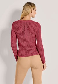 Frau mit langen blonden Haaren, die einen strukturierten rosa Strickpullover und beige Hosen trägt, mit dem Rücken zu einer einfarbigen Hintergrund.