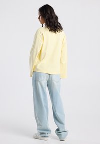 Woman with wavy brown hair wearing a loose pale yellow long-sleeve shirt, light blue wide-leg jeans, and white sneakers, standing against a plain background.