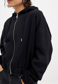 Black zip-up hoodie with a soft texture, featuring a drawstring hood, oversized fit, and ribbed cuffs. Zipper and hem are silver-toned.