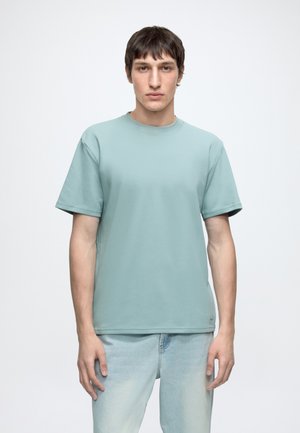 Junger Mann mit kurzen braunen Haaren, der ein schlichtes hellblaues T-Shirt und helle Jeans trägt, steht vor einem weißen Hintergrund.