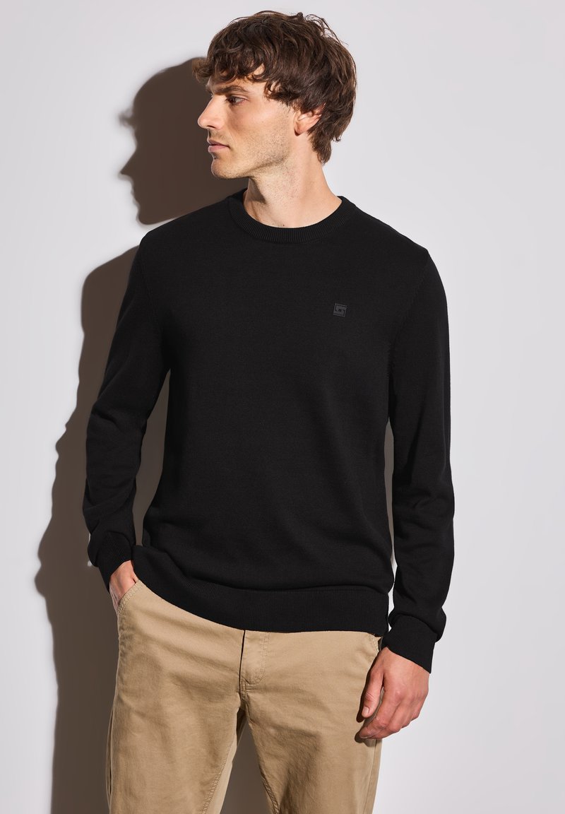 Pull en tricot noir avec un col rond, des poignets et un ourlet côtelés, présentant un petit patch logo sur la poitrine. Porté avec un pantalon beige.