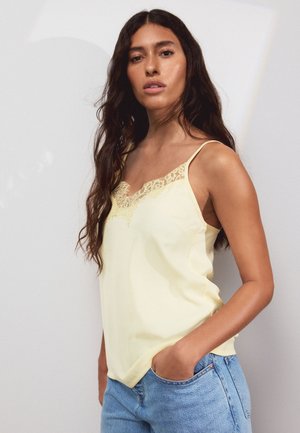 Frau mit langen dunklen Haaren, die eine blassgelbe Spitzen-Camisole und hellblaue Jeans trägt, steht mit einer Hand in der Tasche.
