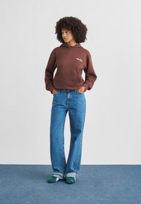 Jeune femme aux cheveux bouclés portant un sweat à capuche marron, un jean large bleu et des chaussures slip-on Puma vertes, debout les mains dans les poches.