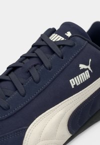 Mørkeblå ruskinds sneakers med en hvid tekstureret stribe og logo. Har flade snørebånd og en polstret krave for komfort. Gummisål er synlig.