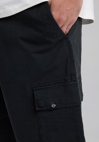Pantalons cargo noirs en tissu texturé, avec une poche latérale et un petit logo. Le design comprend une coupe ample et des coutures renforcées.