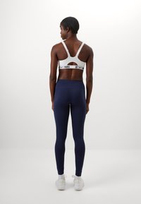 Hvid sports bh med udskæring og sort logo, parret med højtaljede marineblå leggings. Modellen står med ryggen til, iført hvide sko.