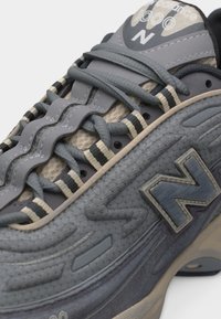 Gros plan sur une chaussure de sport New Balance grise mettant en avant les lacets, la tige texturée et le logo « N » de la marque sur le côté.