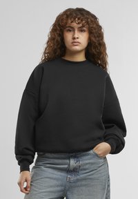 Felpa nera con scollo a girocollo, vestibilità comoda e spalle scese. Il tessuto sembra morbido con polsini a coste, abbinata a jeans di un leggero blu.