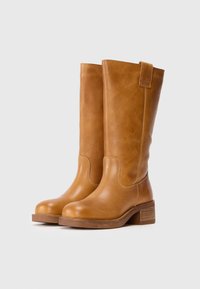 Bottes en cuir beige montantes avec un embout arrondi, des détails cousus et un petit talon en bois. Design sans couture avec une lanière de traction sur le côté.
