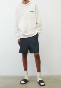 Sudadera beige claro con texto verde, combinada con pantalones cortos azul marino. Calcetines blancos y sandalias negras de dedo al aire completan el look. Diseño de atuendo casual.