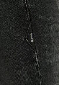 Gros plan sur une poche en denim noir avec des coutures apparentes et une petite étiquette portant l'inscription "CHASIN’" en texte blanc.