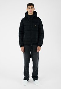 Giacca a vento nera con zip frontale, cappuccio attaccato e polsini elasticizzati. Presenta una tasca sul petto e tasche laterali con zip. Indossata con jeans scuri.