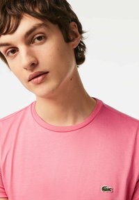 Rosa bomullströja med rund halsringning och en liten grön krokodil-logotyp på vänster bröst. Minimalistisk och slät textur.