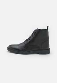Botas de tobillo de cuero negro con un acabado texturizado, punta redonda, diseño con cordones y una suela de goma baja. Incluye una pestaña en la parte posterior.