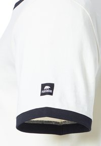 Camisa blanca de manga corta con cuello y dobladillos de las mangas en azul marino. Cuenta con un parche de logo negro en la manga derecha con un diseño de oso polar.