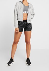 Šedá zipová mikina, šedá sportovní podprsenka s bílým logem, černé šortky s abstraktním vzorem a černé sportovní boty.