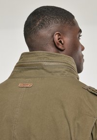 Olive green jacket with a high collar, visible metal zipper, and a leather accent tag. Smooth texture and structured design.