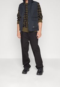 Svart vadderad väst över en olivgrön hounds-tooth tröja, i kombination med svarta byxor och svarta skor. Vesten har en framficka.