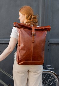 Rödhårig kvinna i ljusa kläder står bredvid en cykel och håller en stor brun läderryggsäck med rullöppning mot en mörk vägg.