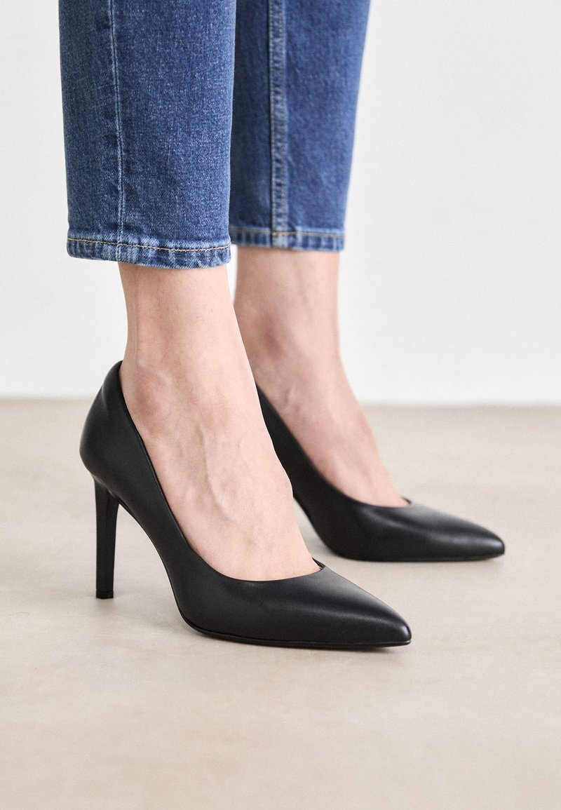 Pieds chaussés d'un jean bleu ajusté et de talons hauts noirs à bout pointu, posés sur un sol clair devant un fond uni.