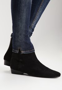 Botas de ante negras hasta el tobillo con punta afilada y acentos en forma de estrella en el costado. Combinadas con jeans de mezclilla oscuros, que destacan un diseño ajustado.