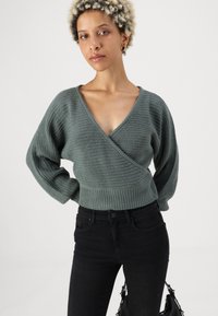 Green wrap sweater with a deep V-neck, textured knit, and wide sleeves. Paired with black jeans and a black handbag.