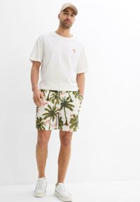 T-shirt blanche à manches courtes avec un petit logo orange en forme de palmier, associée à un short blanc et vert à motifs de palmiers, et des baskets blanches.