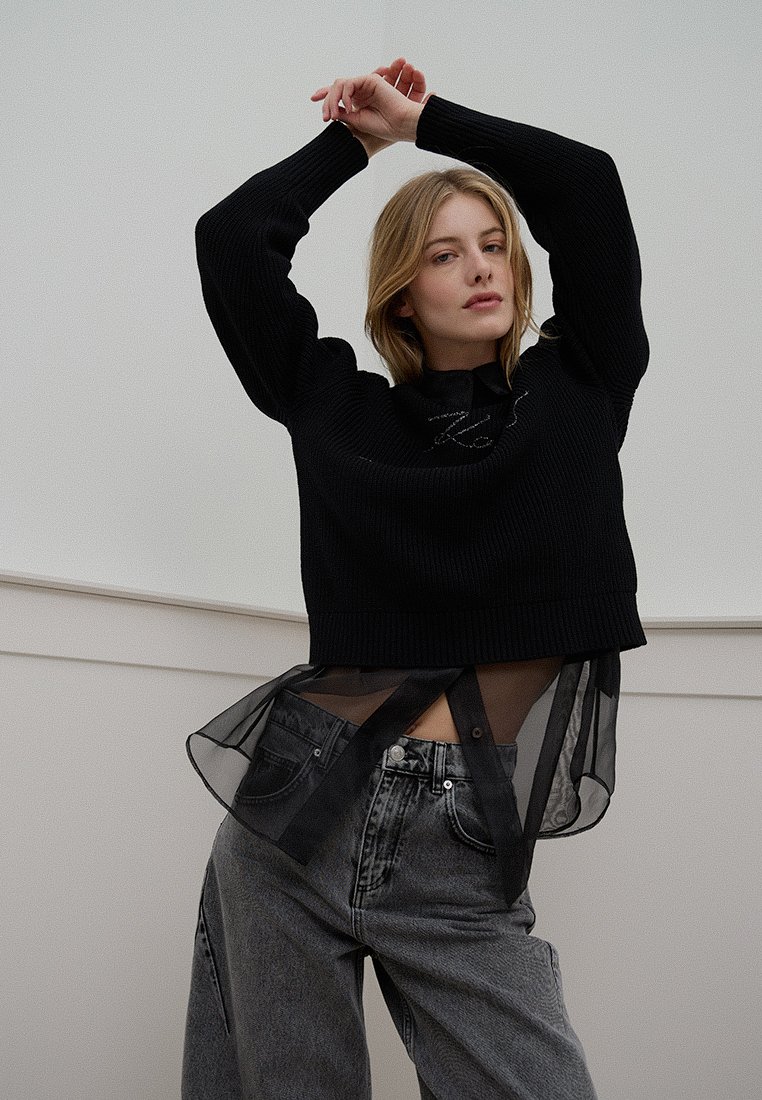 Pull en maille noire à côtes avec une coupe courte, superposé à une chemise en maille noire transparente ; associé à un pantalon en denim gris taille haute.