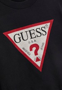 Zwarte sweatshirt met een driehoekig rood en wit logo met rhinestones, waarop "GUESS" en een gestileerd vraagteken in het midden staan.