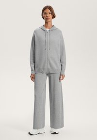 Siva zip-up kapuca s vezicami, v kombinaciji s širokimi sivimi hlačami. Mehka, teksturirana tkanina s sproščenim prileganjem in rebrastimi detajli. Bele superge.