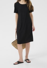 Schwarzes kurzärmeliges Kleid mit rundem Ausschnitt, knielang, mit zwei seitlichen Taschen. Kombiniert mit einer gewebten Strohtasche und flachen schwarzen Sandalen.