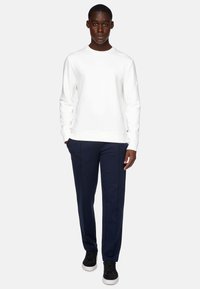 Felpa bianca con polsini e orlo a coste, abbinata a pantaloni blu scuro dal taglio slim. Il modello indossa sneaker nere.