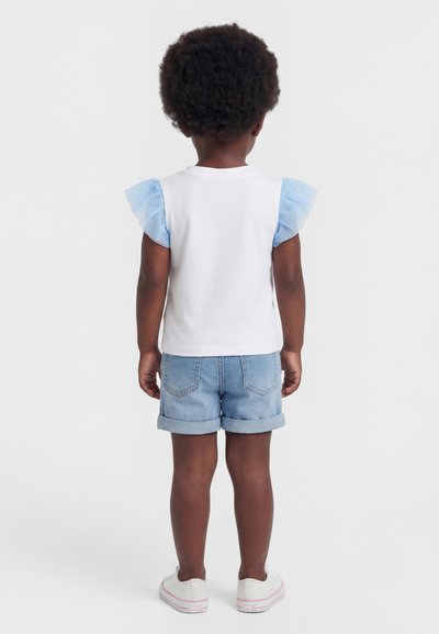 Enfant debout, dos tourné, portant une chemise blanche avec des manches à volants bleus, un short en jean clair et des baskets blanches.