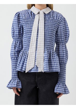 Vrouw met een blauw geruite gesmockte blouse met pofmouwen en een contrasterende licht geruite lange dassenvormige kraag, gecombineerd met een zwarte broek.