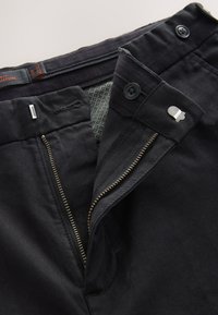 Pantalon noir avec fermeture éclair, bouton et tissu lisse. L'intérieur présente une doublure texturée avec un motif subtil.