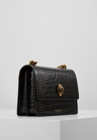 Černá kabelka z kůže s krokodýlím vzorem, zlatým řetízkovým popruhem a zapínáním ve tvaru mušle. Má pevný tvar a ploché zavírání.