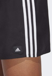 Pantaloni sportivi neri con tre strisce verticali bianche sui lati. Presentano un'etichetta con il logo Adidas sull'orlo. Tessuto liscio.