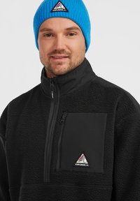 Pull en polaire noir avec une poche zippée sur la poitrine, présentant une finition texturée et un logo triangulaire coloré. Associé à un bonnet en tricot bleu.