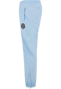 Light blue jogger pants with an elastic waistband, side pockets, and a black circular logo patch on the thigh. Tapered leg design.