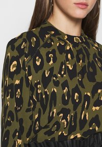 Top verde con stampa leopardata in nero e beige. Il tessuto ha una consistenza morbida con un design plissettato sul colletto e dettagli neri.