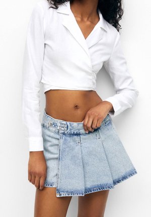 Vrouw draagt een wit cropped wikkel-top met lange mouwen en een lichtblauwe geplooide denim minirok, staand tegen een witte achtergrond.