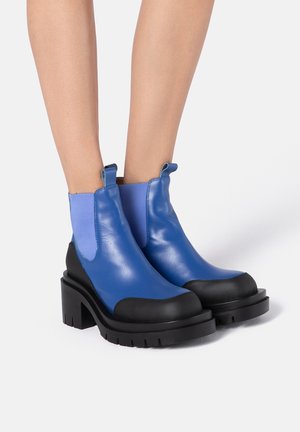 Niebieskie botki z błyszczącym wykończeniem ze skóry, z czarnymi gumowymi podeszwami i elastycznymi panelami po bokach. Kwadratowy nosek z podniesionym obcasem.