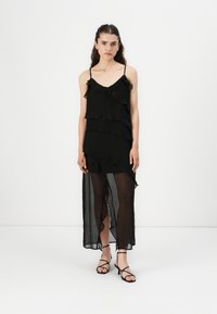 Robe noire avec des volants superposés, bretelles spaghetti et une partie inférieure transparente. Comprend une fente haute et des sandales plates noires.