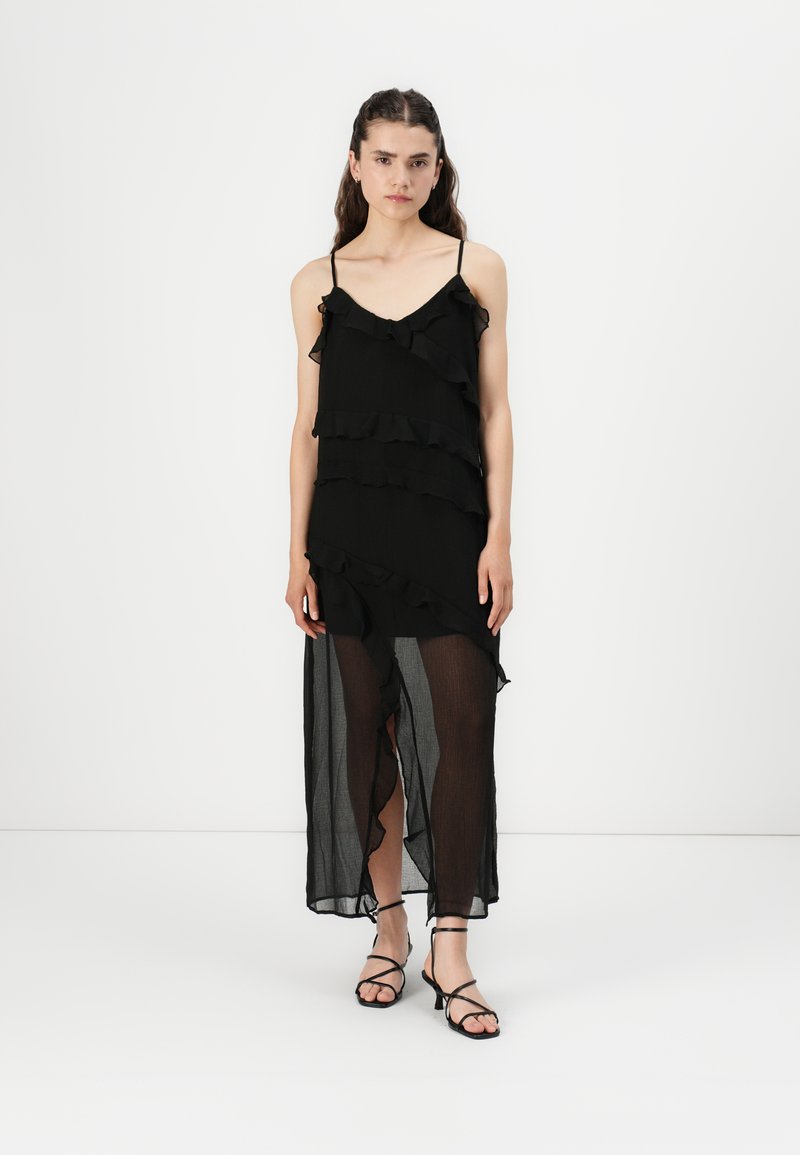 Robe noire avec des volants superposés, bretelles spaghetti et une partie inférieure transparente. Comprend une fente haute et des sandales plates noires.